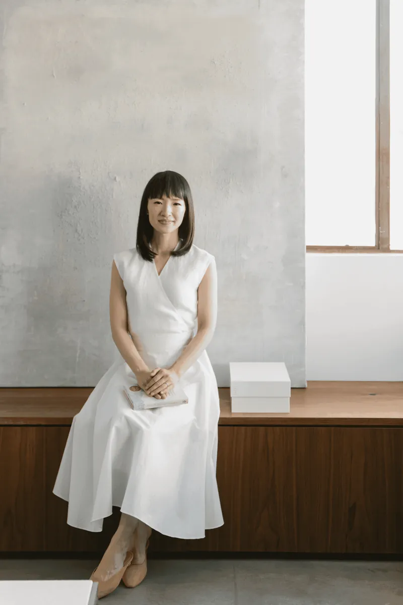 Marie Kondo seated on a wooden bench, holding her book